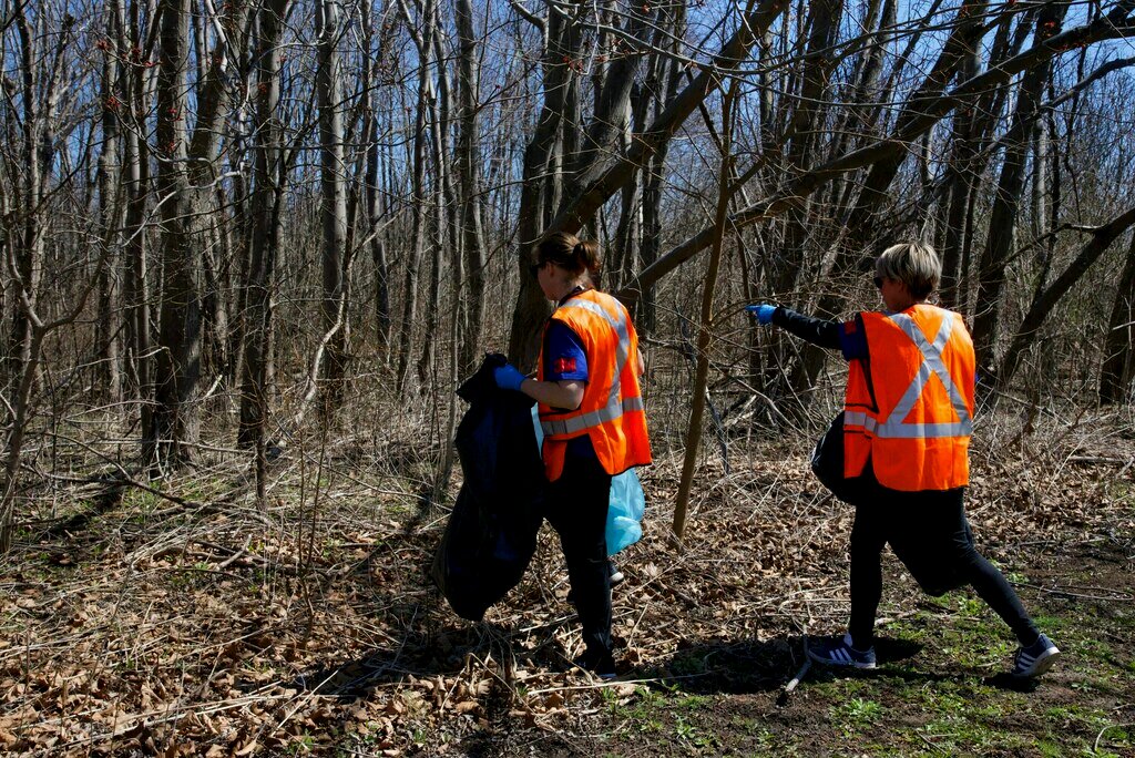 Two 3Mers wearing blue 3M volunteer shirts, orange high visibility vests, blue gloves, and hold garbage bags.